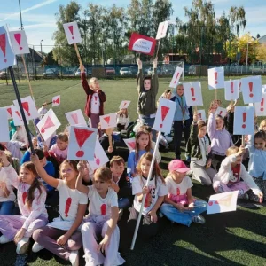 Grupa uczniów na boisku unosi białe plakaty z czerwonymi wykrzyknikami.